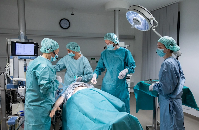 Surgical team in scrubs performing procedure on patient in operating room with vital signs monitor and sterile equipment.