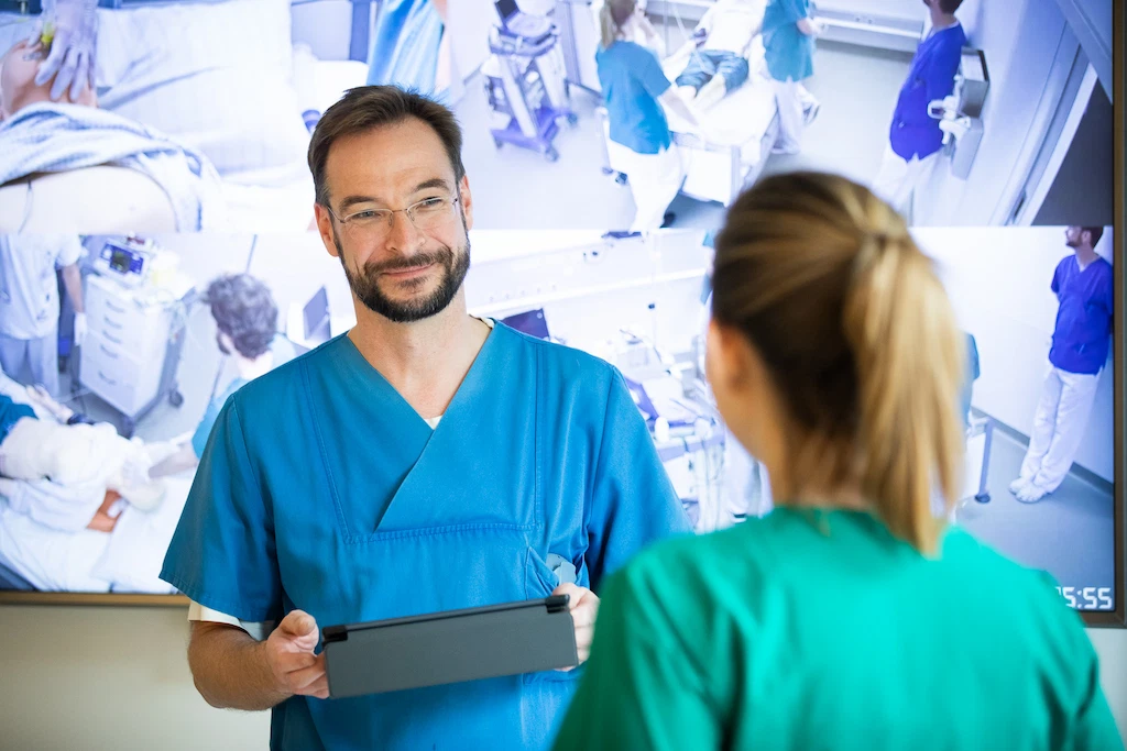 Instructor using a control tablet next to a training participant during a video debriefing session, with the debriefing screen visible in the background.