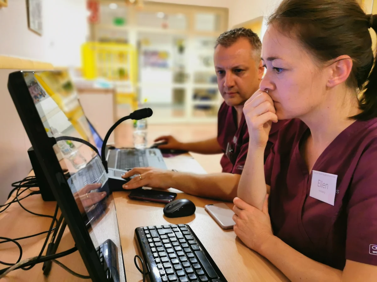 Two medical professionals, Ellen and a colleague, intensely monitoring a live simulation on a computer screen while using a SIMStation recording and communication interface.