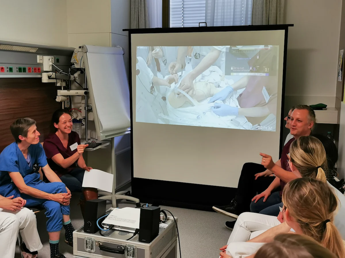 A medical team participating in a post-simulation debriefing session, watching a projected video playback of a neonatal resuscitation exercise to analyze team communication and clinical performance.
