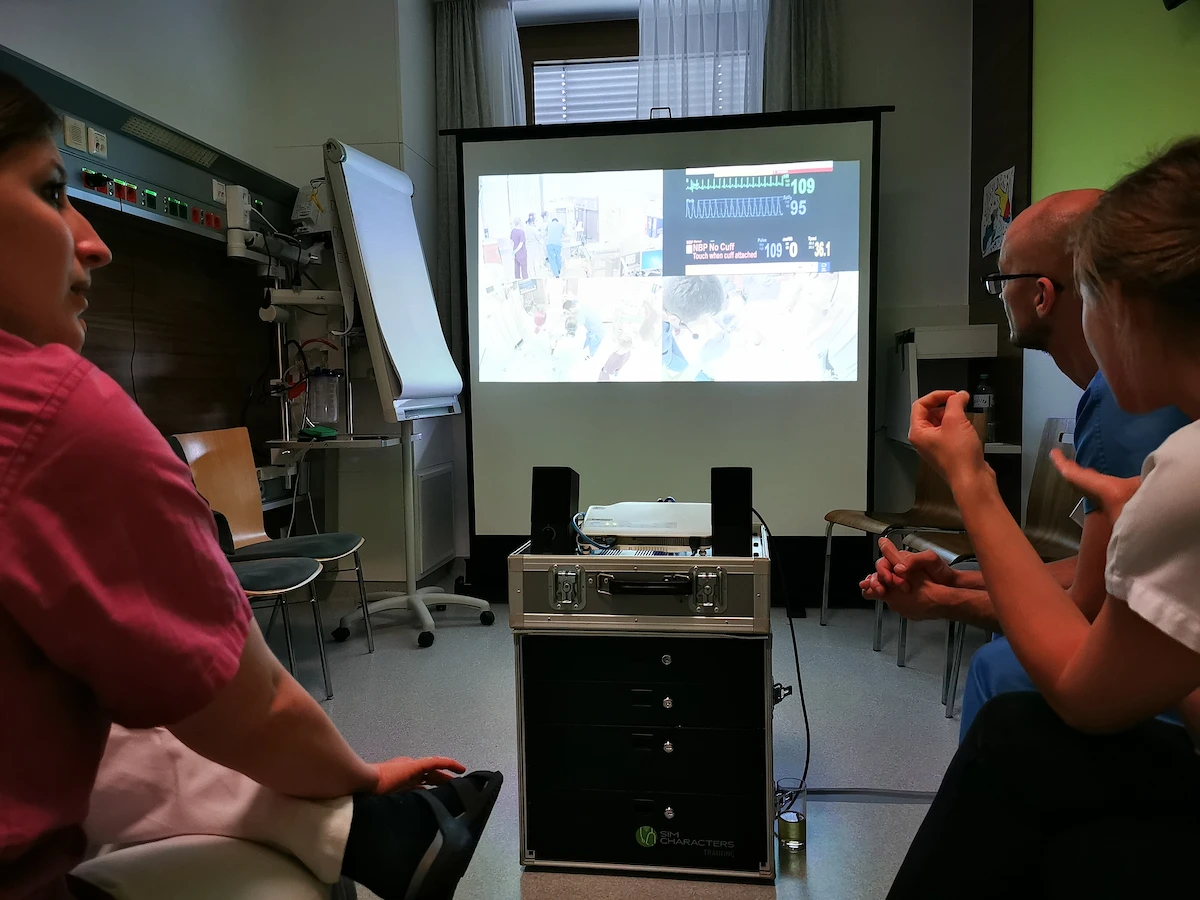 A clinical debriefing session where a healthcare team uses a mobile SIMStation unit to project and analyze multi-view video and vitals data from a completed medical simulation exercise.