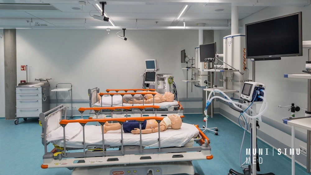 A modern clinical simulation ward at MUNI SIMU MED featuring multiple patient manikins on mobile hospital beds, equipped with SIMStation ceiling-mounted cameras and bedside monitoring technology for medical training.