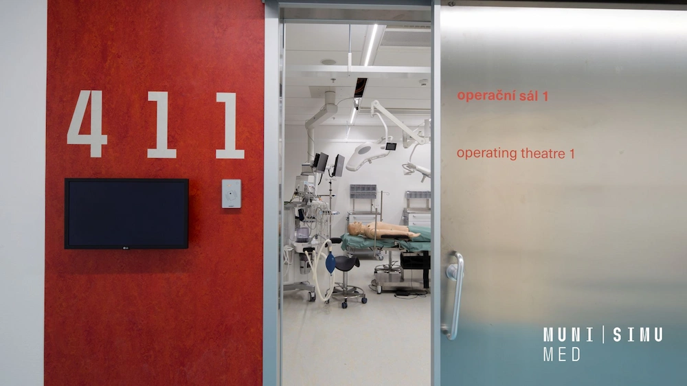 View through an open sliding door labeled 'Operating Theatre 1' at the MUNI SIMU MED facility, showing a realistic surgical simulation room with a patient manikin, surgical lighting, and integrated medical monitoring equipment.