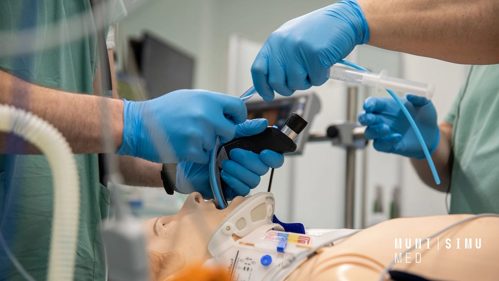 A close-up of medical trainees in blue gloves performing a precision intubation on a high-fidelity patient manikin wearing a cervical collar at the MUNI SIMU MED training facility.