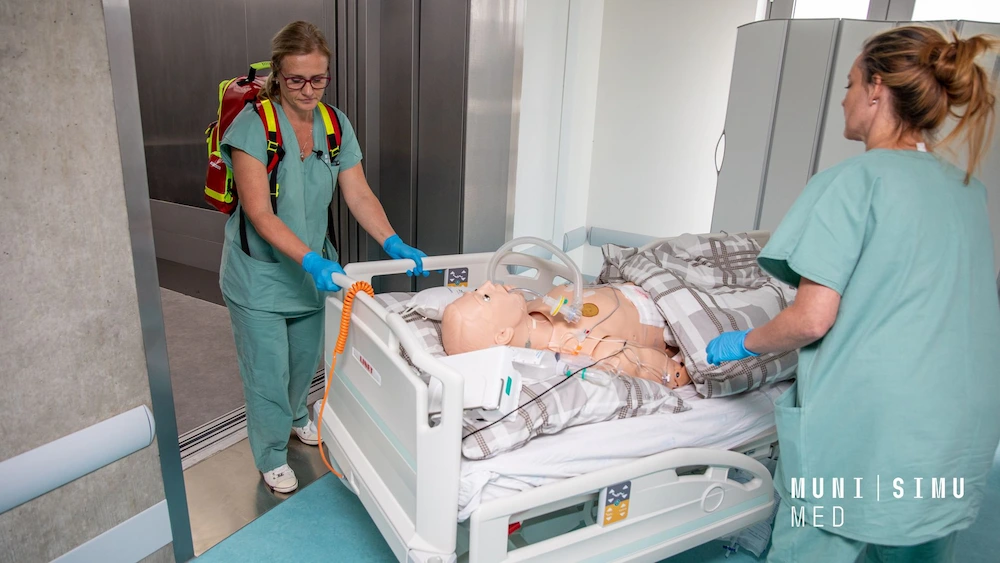 Two medical professionals in teal scrubs at MUNI SIMU MED transport a high-fidelity patient manikin on a mobile hospital bed during a realistic patient transfer simulation.