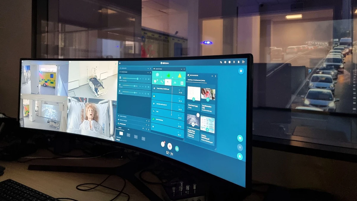 A wide curved monitor displays surveillance feeds, medical data, and control panels in a dimly lit room. With the SIMStation Training Suite and video debriefing, the platform keeps you fully in command during professional daily simulation practice.