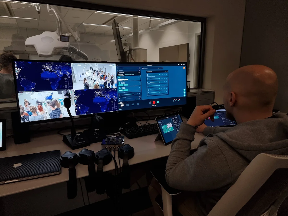 A person sits at a control station with multiple monitors displaying a world map, a video call with several people, and various control panels for evaluation and video debriefing in a modern, dimly lit room.