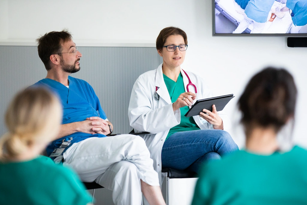 Two medical professionals, one in blue scrubs and one in a white coat with a stethoscope, sit and talk while looking at a tablet—evaluating performance with the SIMStation Training Suite and video debriefing. Two others in green scrubs face them.