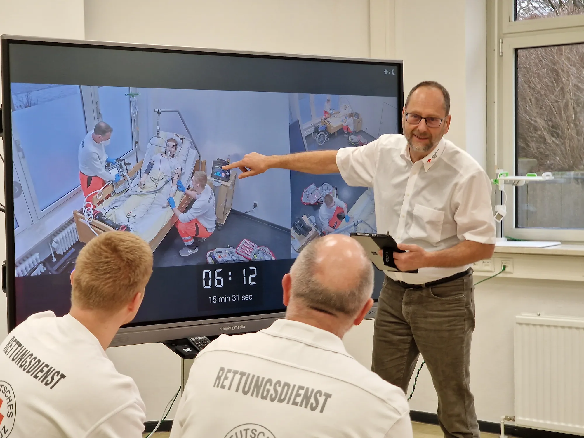 A man points at a large screen showing a medical simulation with paramedics attending to a patient, while two people in “Rettungsdienst” uniforms watch and listen—recording and evaluation ensure your training meets professional standards.